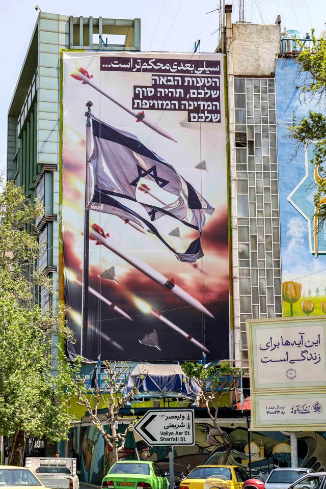 Sebuah spanduk bergambar rudal dan drone terbang melewati bendera Israel yang robek, dengan teks dalam bahasa Persia di Teheran, Jumat (19/4/2024) Foto: ATTA KENARE / AFP