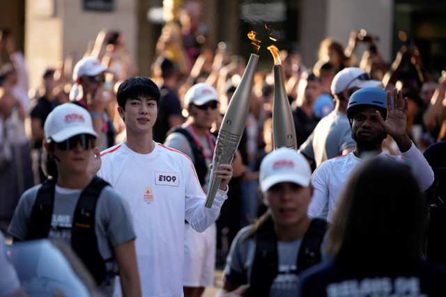 Jin BTS membawa obor Olimpiade 2024 di area Museum Louvre, Paris, Minggu (14/7/2024). Foto: Louise Delmotte/AP