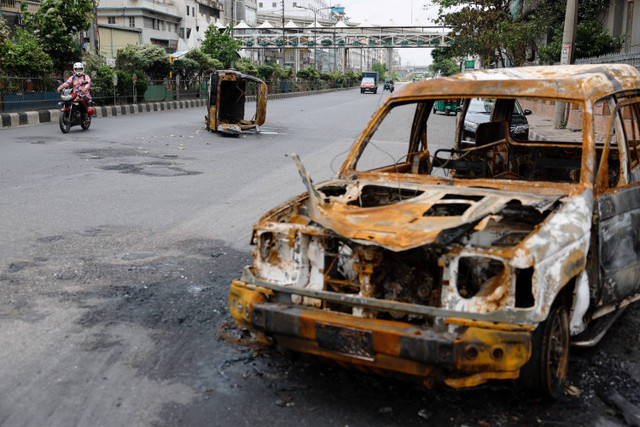 Pengendara motor melewati kendaraan rusak yang dibakar imbas kerusuhan protes terhadap penolakan kebijakan kuota kerja di Dhaka, Bangladesh, Senin (22/7/2024). Foto: Mohammad Ponir Hossain/ REUTERS