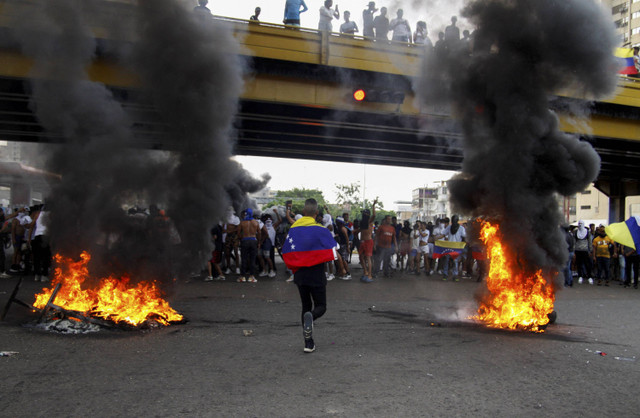 Seorang demonstran berlari dengan membawa bendera Venezuela di antara penghalang jalan yang terbakar selama protes terhadap hasil pemilu presiden di Venezuela, Minggu (29/7/2024). Foto: Samir Aponte/REUTERS