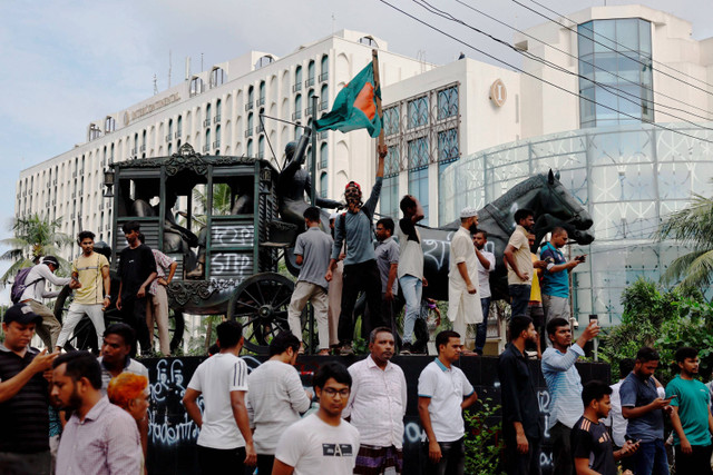 Seorang pria yang memegang bendera Bangladesh berdiri di depan sebuah kendaraan yang dibakar di Ganabhaban, kediaman Perdana Menteri, setelah pengunduran diri PM Sheikh Hasina di Dhaka, Bangladesh, 5 Agustus 2024. Foto: Mohammad Ponir Hossain/Reuters