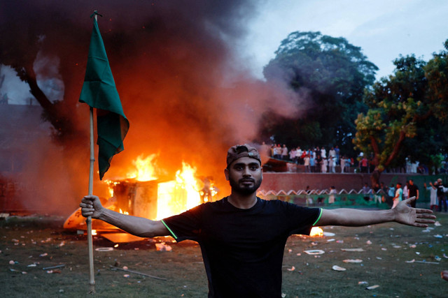 Seorang pria yang memegang bendera Bangladesh berdiri di depan sebuah kendaraan yang dibakar di Ganabhaban, kediaman Perdana Menteri, setelah pengunduran diri PM Sheikh Hasina di Dhaka, Bangladesh, 5 Agustus 2024. Foto: Mohammad Ponir Hossain/Reuters