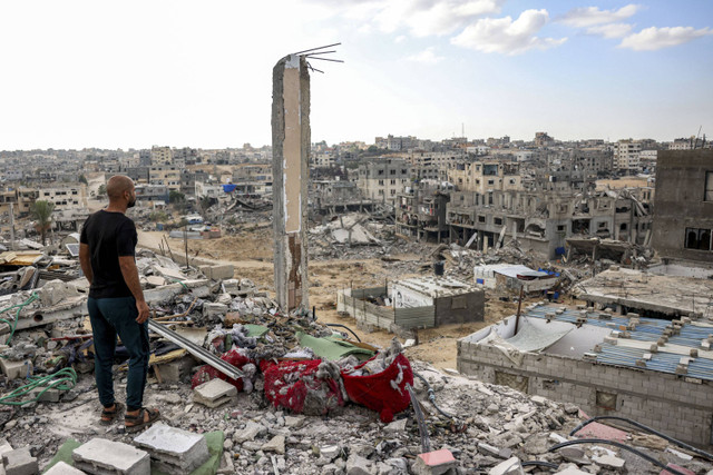 Seorang pria berdiri di atas sebuah bangunan yang rusak parah dan melihat bangunan lain yang hancur di Khan Yunis, Jalur Gaza selatan, Senin (7/10/2024). Foto: Bashar Taleb/AFP