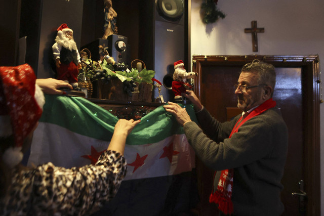 Umat Kristen Suriah menggantungkan bendera era kemerdekaan Suriah saat merayakan malam Natal di rumah mereka di Latakia, Suriah, Selasa (24/12/2024). Foto: Aaref Watad/AFP