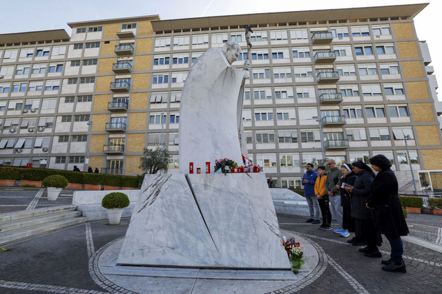 Orang-orang berdoa saat lilin dan bunga diletakkan di dasar patung mendiang Paus Yohanes Paulus II di luar Rumah Sakit Gemelli tempat Paus Fransiskus dirawat, di Roma, Italia, Minggu (23/2/2025). Foto: Ciro De Luca/REUTERS