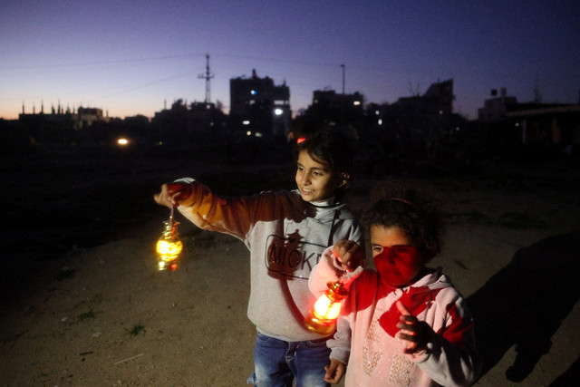 Dua anak Palestina menyalakan lentera tradisional usai berbuka puasa di tengah bangunan yang hancur di kamp pengungsi Bureij, Jalur Gaza utara, Sabtu (2/3/2025). Foto: Eyad BABA / AFP