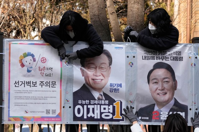 Pejabat dari panitia penyelenggara pemilu memasang poster calon presiden Lee Jae-myung dari Partai Demokrat yang berkuasa dan Yoon Suk Yeol, kanan, dari oposisi utama People Power Party di Seoul, Korea Selatan, 18 Februari 2022. Foto: Ahn Young-joon/AP Photo