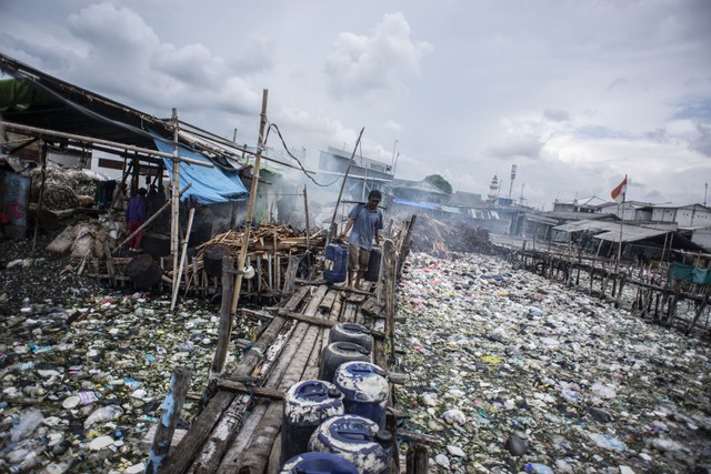 Seorang warga melintasi jembatan kayu di antara tumpukan sampah di kawasan perkampungan nelayan Cilincing, Jakarta, Minggu (20/2/2022). Foto: Aprillio Akbar/ANTARA FOTO