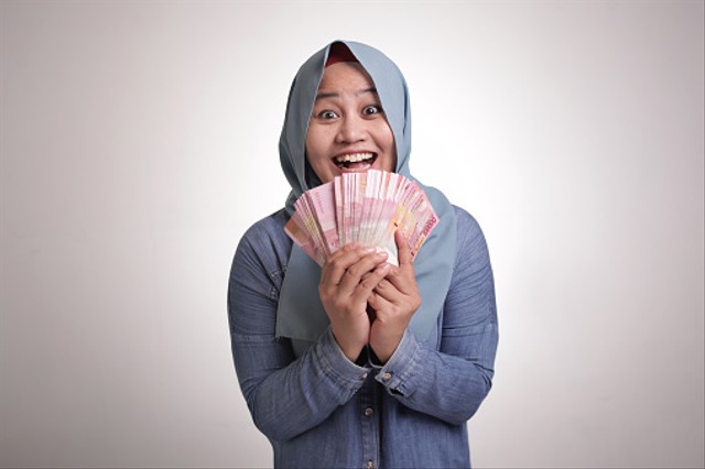 Portrait of Indonesian muslim woman holding rupiah money, smiling laughing winning gesture (Foto diambil dari iStock karya airdone)